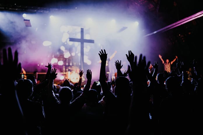 Foto: LIAM SHAW + IA Jóvenes con los brazos en alto durante un concierto, símbolo de búsqueda colectiva y emoción compartida